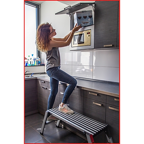 Frau in Jeans und Top steht auf Tritt, um Schrank über Mikrowelle zu öffnen. Küche im Hintergrund.